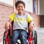 A smiling young boy in a red wheelchair in a hallway.