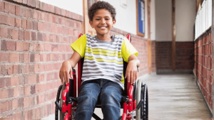A smiling young boy in a red wheelchair in a hallway.